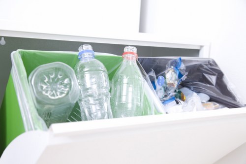 A close-up of an open white waste container or recycling bin filled with rubbish, including clear plastic bottles with blue and red caps, resting inside a bright green inner compartment. Next to the bottles, there is a black plastic bag containing additional waste materials such as plastic packaging or miscellaneous rubbish. The container is situated on a clean, smooth surface, possibly inside an office or utility area, with a white wall or partition visible in the background. The image emphasizes the organized collection of recyclable materials alongside general waste, illustrating a typical rubbish removal setup aligned with services provided by Office Clearance Paddington, serving the Paddington area. The lighting is even, highlighting the details of the bottles’ transparent plastic and the contrasting black bag.