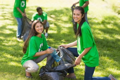 Four young volunteers wearing bright green T-shirts with recycling symbols participate in a community rubbish cleanup on a grassy outdoor area. Two women are in the foreground, smiling as they hold open a large black trash bag. One woman, kneeling on the left, has long brown hair and is wearing beige trousers; she is holding a plastic bottle and appears to be ready to dispose of it properly. The other woman, standing on the right, has long dark hair tied back and is wearing dark blue jeans, actively assisting with the trash collection. In the background, two additional volunteers can be seen, one holding the top of a trash bag and the other bent over, both involved in the cleanup effort. They are surrounded by sunlight filtering through trees, creating an engaging and positive atmosphere typical of community environmental initiatives. This scene reflects volunteering for rubbish collection, possibly supported by local waste management services such as Office Clearance Paddington, highlighting the importance of community involvement in maintaining clean public spaces in the Paddington area, relevant to local environmental efforts and waste disposal practices.