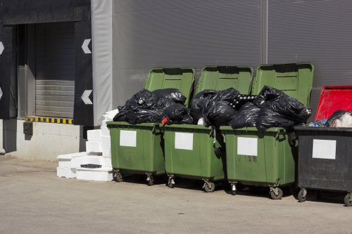 In the foreground, three green wheeled bins with open lids are filled with black rubbish bags, placed on a concrete surface outside a commercial building. The rubbish bags appear to be full and loosely packed, with some bags slightly bulging and tied at the top. To the right of the green bins, a black bin with a white label is also filled with rubbish, and a red bin is partially visible. Behind the bins, there is a grey industrial wall with a roller shutter door that is partially open, revealing a dark interior. Adjacent to the shutter, a set of white bricks is stacked on the ground, near the red bin. The scene is well-lit with natural daylight, indicating an outdoor setting in Paddington, London, consistent with local waste management operations for office clearance and rubbish disposal services provided by Office Clearance Paddington.