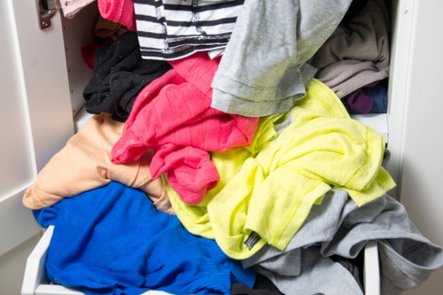 A close-up view of an open metal filing cabinet drawer filled with a disorganized assortment of crumpled and folded clothing items, including brightly colored textiles such as red, yellow, blue, and grey fabric, with some pieces spilling over the edges. The surrounding environment suggests an indoor setting, possibly an office or storage area, with visible white shelving or cabinet parts in the background. This cluttered scene reflects rubbish or waste materials typically associated with clearance or disposal services, similar to those offered by Office Clearance Paddington, serving the postcode area near Westminster and Paddington in London.
