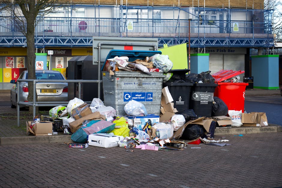 A man with curly black hair, wearing a dark grey t-shirt and grey plaid shorts, is standing outdoors on a paved surface next to a large white skip or waste container filled with disorganized burnt-ora