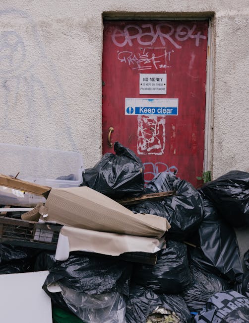 A cluttered scene outside a building featuring a collection of black rubbish bags piled against a textured, off-white wall with visible wear. The rubbish bags are filled and tied, with some appearing