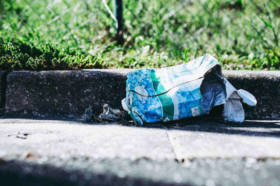 A cluttered scene outside a building featuring a collection of black rubbish bags piled against a textured, off-white wall with visible wear. The rubbish bags are filled and tied, with some appearing
