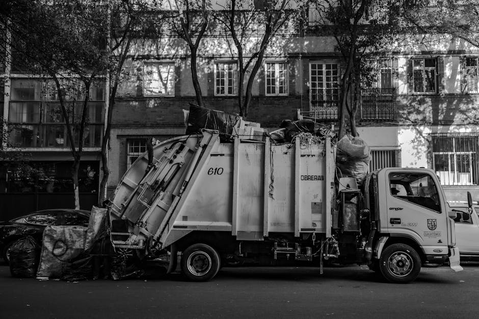 A large blue wheeled rubbish bin made of durable plastic with a textured surface, positioned upright on a paved surface next to a white delivery van with a metal door. The bin's lid is closed, and it