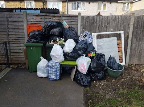 A collection of black, white, and blue patterned rubbish bags piled on and around a small green garden bench in a residential outdoor area, with some bags leaning against a wooden fence made of vertical planks. To the left, a green wheelie bin and an orange recycling bin are visible, with additional black bags stacked on top of the recycling bin. Behind the bags, there is an old, disused white bedframe or headboard leaning against the fence, along with a plastic storage container. The ground features a paved section and a patch of soil with sparse grass, indicating an urban backyard setting. The scene appears to be part of a rubbish removal effort undertaken by Office Clearance Paddington, possibly in the Paddington area, highlighting waste collection and clearing activities suitable for local rubbish disposal services.