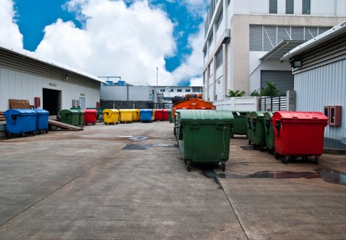 A spacious outdoor area situated between modern multi-storey office buildings, featuring a wide concrete ground surface with several large, mobile wheelie bins arranged in a semi-organised line. The bins are made of durable plastic with varying colours including green, red, yellow, and blue, indicating different waste types. On the left side, two white industrial storage units with open doorways are visible, while the background showcases additional warehouse-like structures and a partly cloudy sky with patches of blue. The scene appears to be part of a commercial property in Paddington, used by Office Clearance Paddington for rubbish collection and disposal. The environment is clean and well-maintained, with the bins positioned for easy access, and the overall setting reflects typical rubbish management operations in an urban office district.