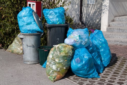 A collection of various rubbish bins and bags positioned on a paved pavement area, with some bins being large plastic or metal containers and others being tightly tied blue and yellow plastic bags filled with waste. The bags contain visible mixed rubbish, including foliage or plant material, and are placed next to a concrete wall with stairs leading up behind them. The scene appears to be part of an outdoor rubbish collection point, with some greenery visible in the background, suggesting a residential or commercial area within Paddington. The arrangement indicates an outdoor waste collection zone managed by Office Clearance Paddington, specialising in rubbish removal services for property clearances in the local postcode area. The lighting appears natural, with shadows cast on the ground, emphasizing the outdoor setting and the materials' textures and colours.